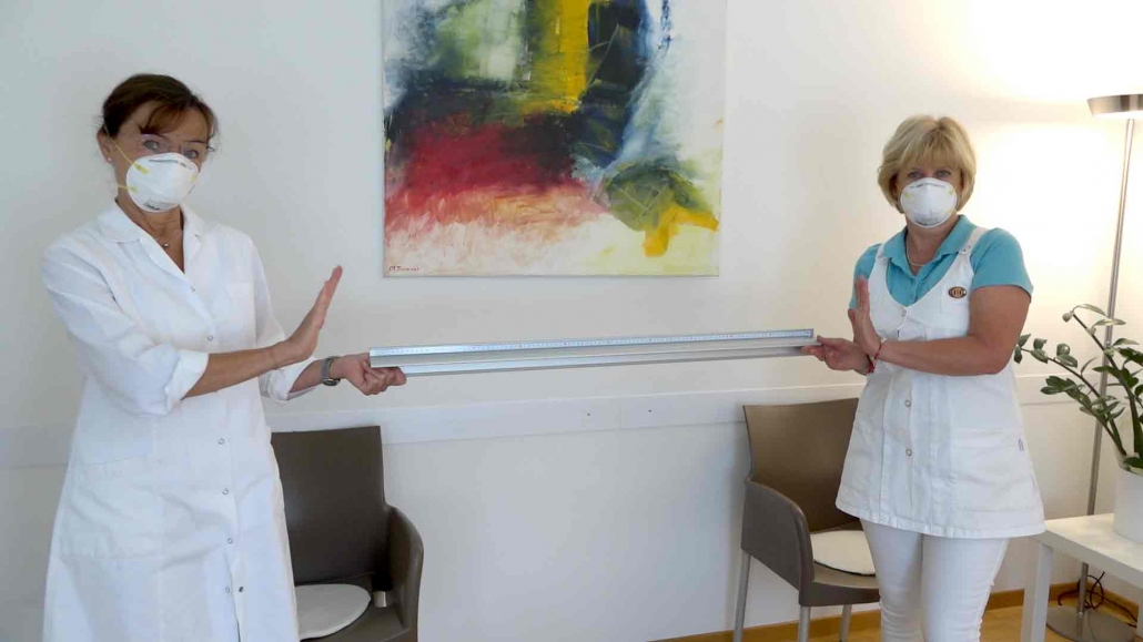 Das Foto zeigt Frau Doktor Astrid Nader mit Ihrer Assistentin im Wartezimmer Ihrer Ordination mit einem Lineal, welches das Abstandhalten veranschaulichen soll.